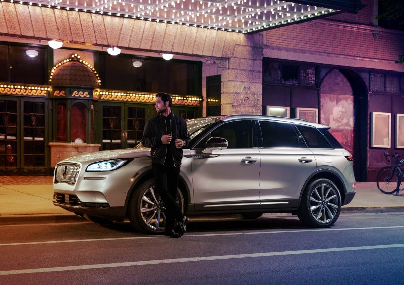 A 2022 Lincoln Corsair SUV is parked outside a theater as the driver relaxes against the frame and lights illuminate the floating roofline and body | Loveland Lincoln in Loveland CO