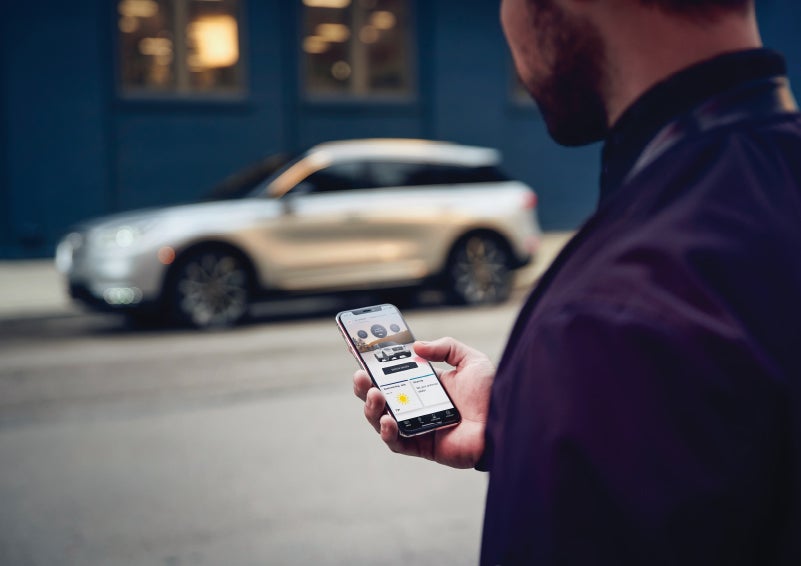 A man is holding his phone looking at the Lincoln Way™ App and we see his 2022 Lincoln Corsair SUV in the background against a blue brick wall | Loveland Lincoln in Loveland CO