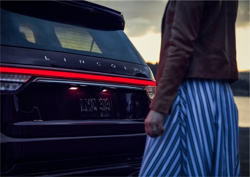 A person is shown near the rear of a 2023 Lincoln Aviator® SUV as the Lincoln Embrace illuminates the rear lights | Loveland Lincoln in Loveland CO