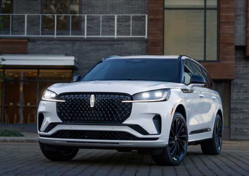 A white 2025 Lincoln Aviator® SUV is shown parked outside a modern building with gray brick and wood accents. | Loveland Lincoln in Loveland CO